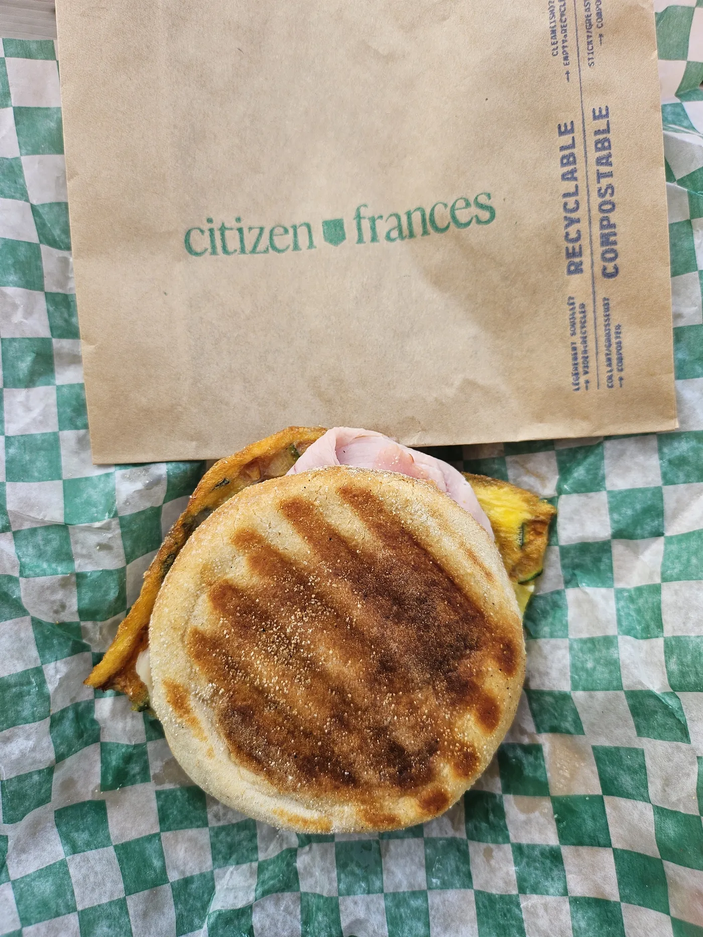A grilled breakfast sandwich on green checked paper, next to a citizen frances paper bag